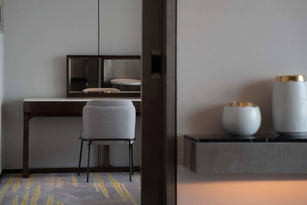 A modern hotel room with a wooden desk, a grey upholstered chair, and a small table with two decorative vases.