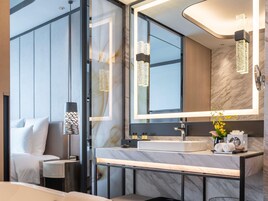 A modern bathroom with a large mirror, marble countertop, and a sink. There is a bedside table with a lamp and a bed with white bedding.