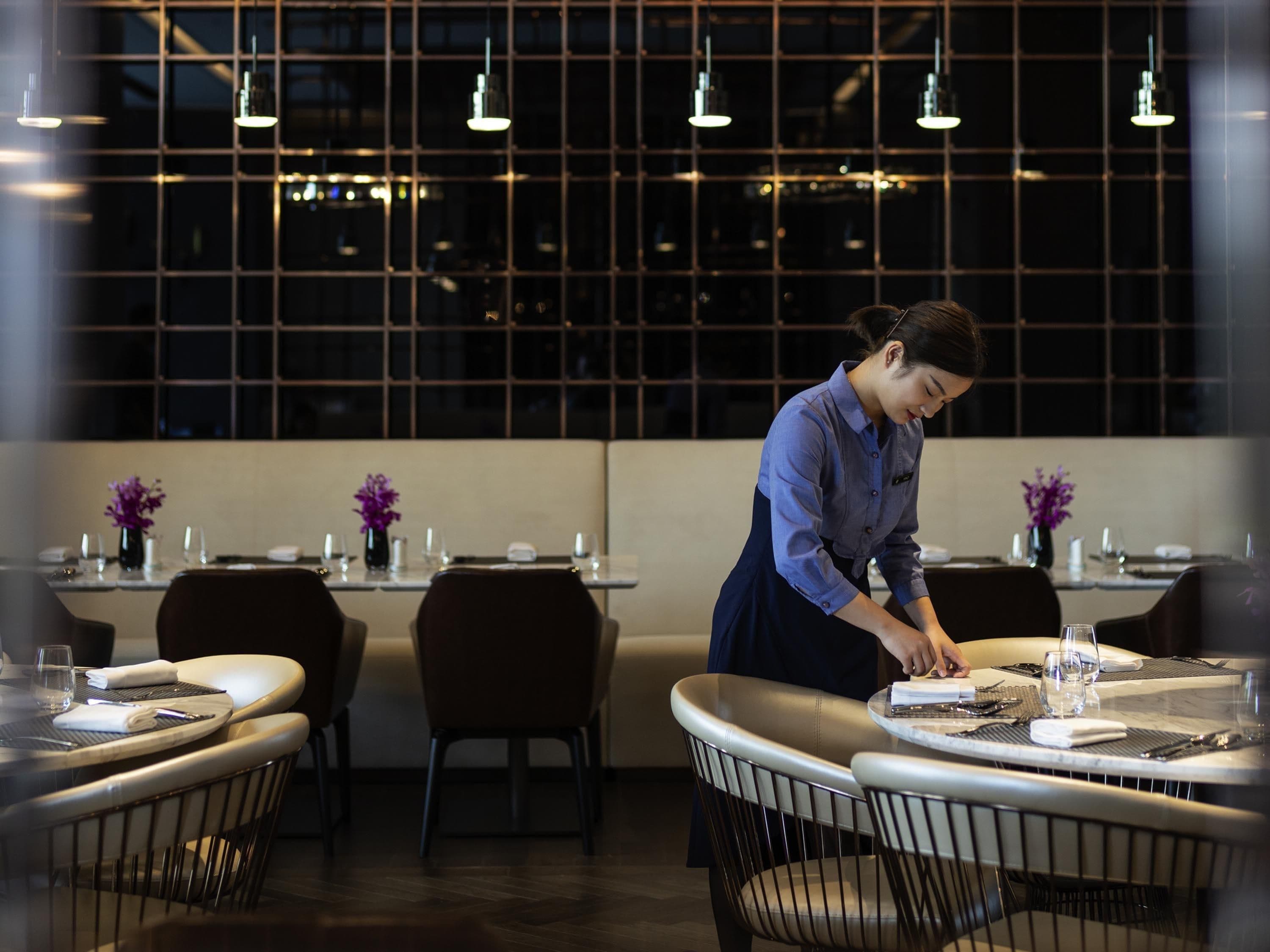 A restaurant with tables set for dining, a waiter attending to a table, and a modern interior design with pendant lighting.