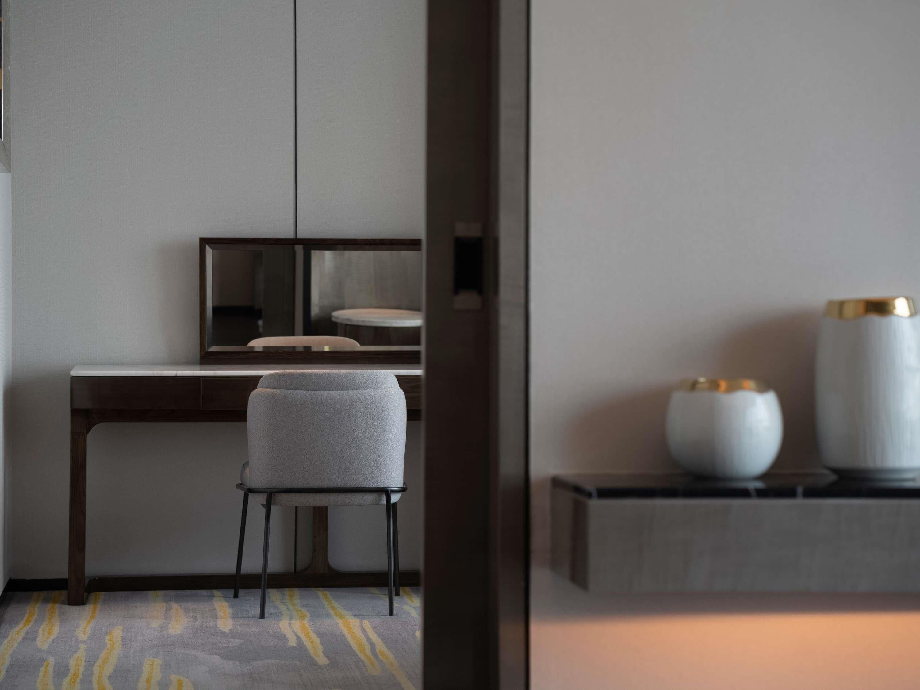 Una habitación de hotel moderna con un escritorio de madera, una silla tapizada de gris y una pequeña mesa con dos jarrones decorativos.