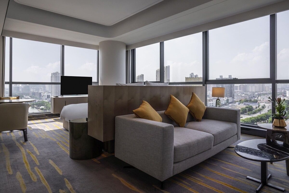 A modern hotel room with a sofa, armchair, and a TV. Large windows offer a city view.