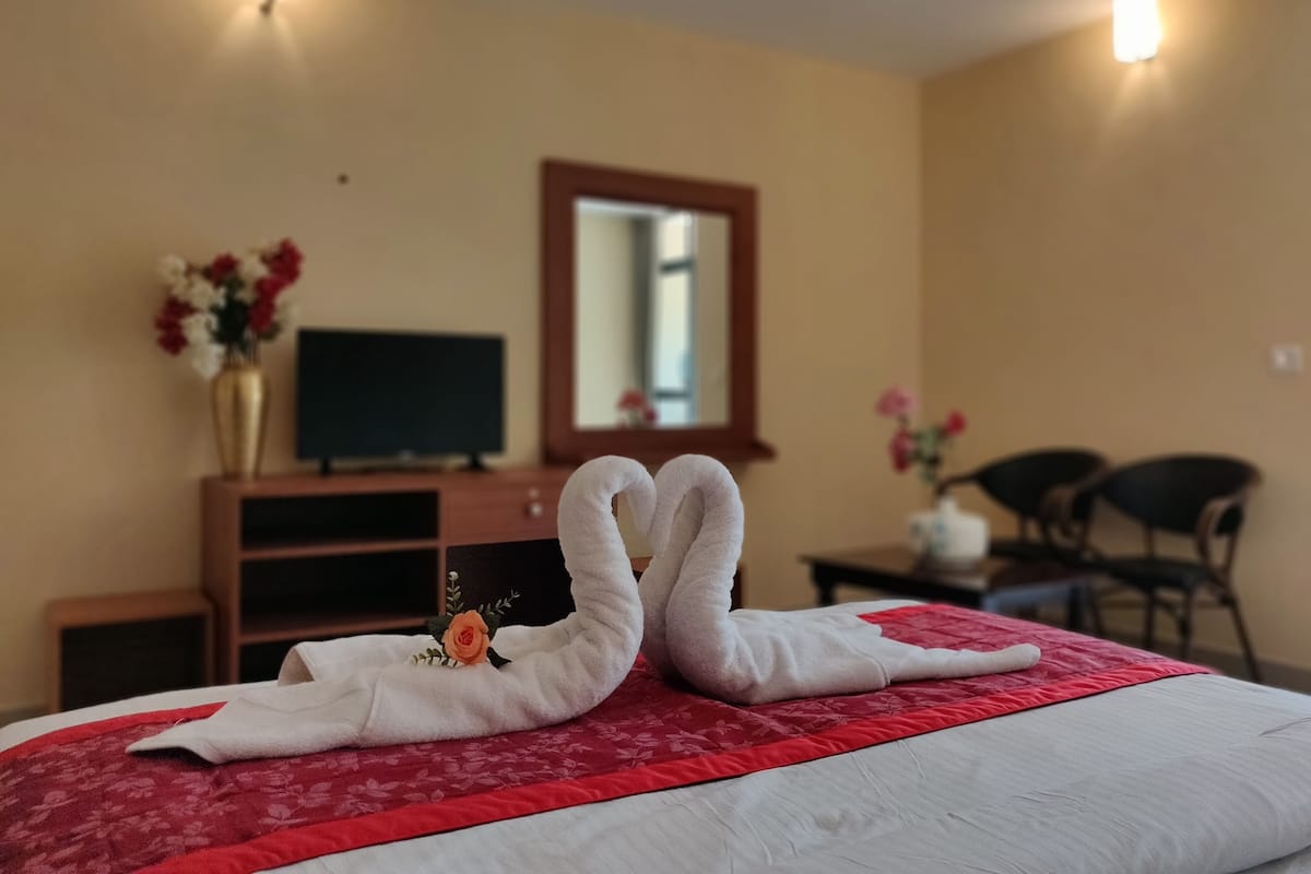 Una habitación de hotel con cama, televisor, espejo y escritorio con un florero.