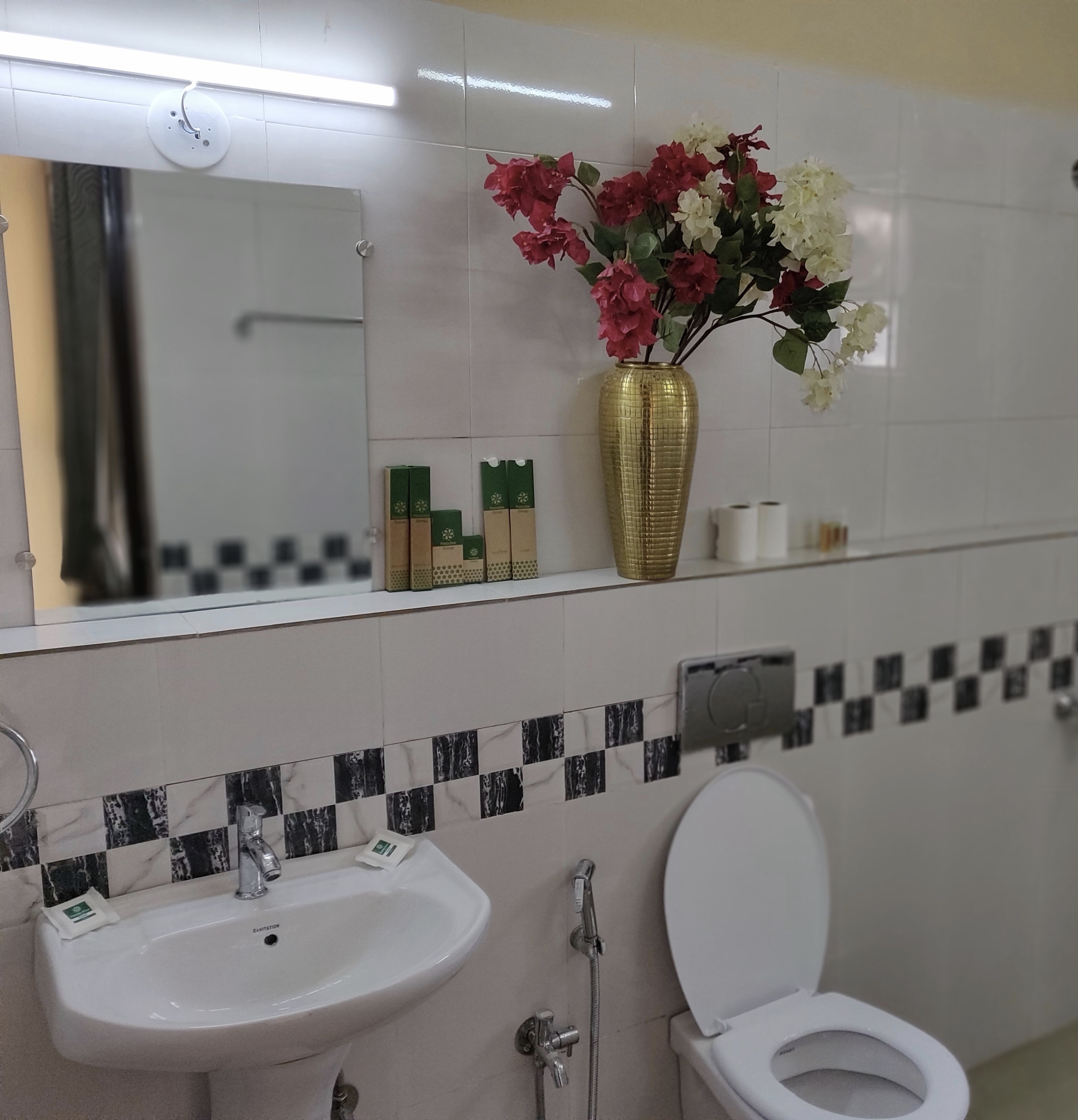 A bathroom with a white sink, toilet, and a mirror above. There is a vase with flowers on the wall and a shelf with toiletries.