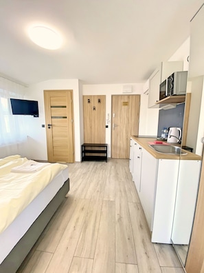 A modern hotel room with a bed, a small bench, a kitchenette, and a TV mounted on the wall.
