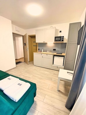 A compact hotel room with a kitchenette, a bed with green bedding, a small table, and a curtain.
