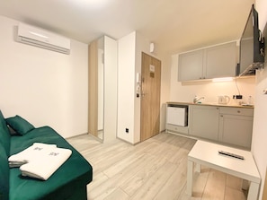 A compact room with a green sofa, a small table, a kitchenette, and a door leading to another room.