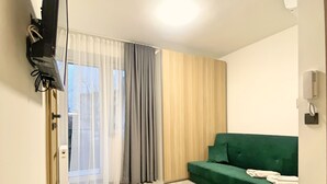 Un soggiorno moderno con un divano verde, una televisione a schermo piatto montata sulla parete, un tavolino con un telecomando e una porta vetrata scorrevole che dà su un balcone.