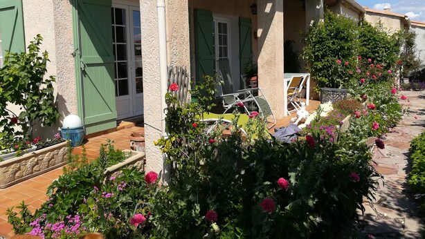 Een gebouw met een overdekte veranda, groene luiken en een tuin met verschillende bloemen.