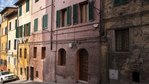 Eine Reihe traditioneller Gebäude mit rundbogigen Durchgängen und grünen Fensterläden, ein weißes Auto parkt auf der Straße, und ein klarer Himmel.