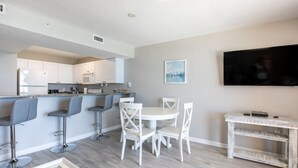 Una cocina moderna con armarios blancos, un microondas y un horno empotrado. Hay una zona de comedor con sillas blancas y una mesa, y un televisor montado en la pared.