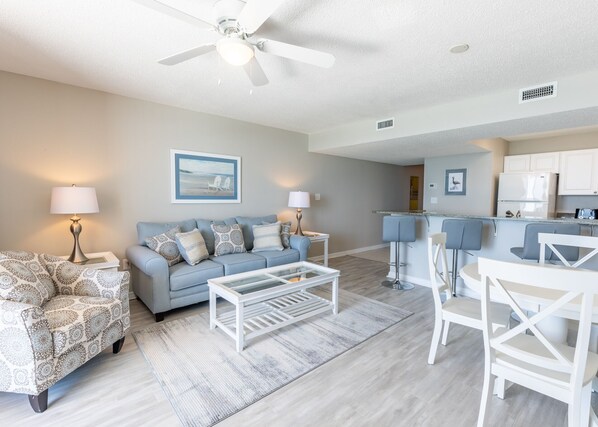 A modern living room with a blue sofa, patterned armchair, and white coffee table. A kitchen area is visible in the background with white cabinets and a bar counter. - Beachfront 1BR in Destin! Balcony, 3 Pools, Hot Tub, Tennis, Steps to Beach. (Destin)