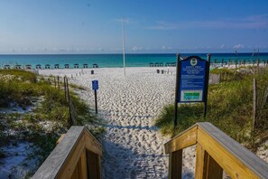 A beach with a wooden walkway leading to the water, a sign with safety instructions, and beach umbrellas in the background.