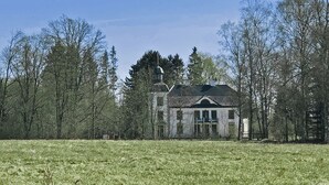 Une maison rustique à deux étages, dotée d’un toit en pente, entourée d’un champ verdoyant et d’une forêt dense.