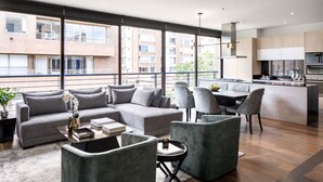 A modern apartment with a living area featuring a grey sectional sofa, a small round table, and two grey armchairs. There is a kitchen area with a dining table and chairs, and large windows offering views of the city. -  ATTIK SUPERIOR -501 (Bogotá)