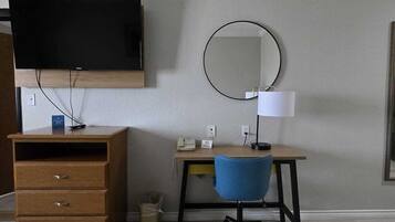 Una habitación con una televisión de pantalla plana montada en la pared, un escritorio de madera con una silla, un espejo redondo y un cesto de basura pequeño.
