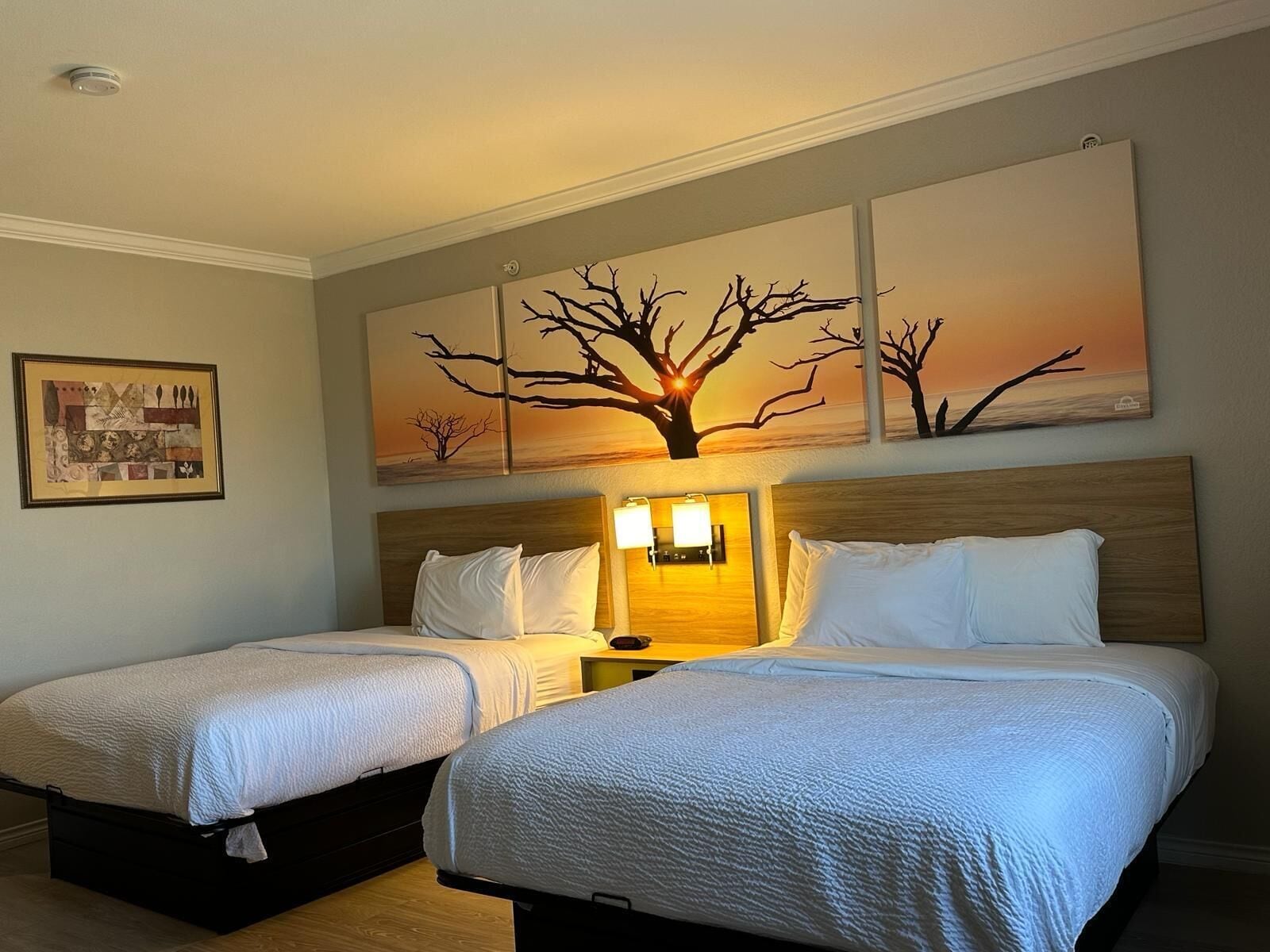 Habitación de hotel con dos camas, una mesita de noche con lámparas y un cuadro grande con un paisaje de árboles.