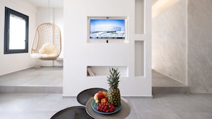 A modern interior with a TV mounted on a white wall, a hanging chair, and a fruit bowl on a table. - George & Joanna Suites (Santorini)