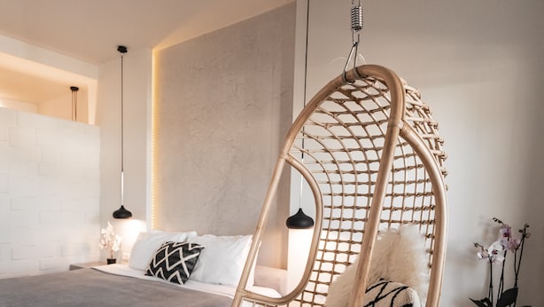 A modern bedroom with a hanging chair, a bed with white and black pillows, a bedside table, and a potted plant. - George & Joanna Suites (Santorini)