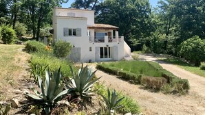 Una casa de dues plantes amb un balcó, envoltada de vegetació i un camí de terra.