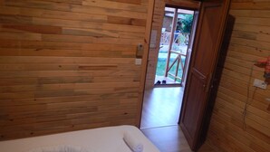 A wooden room with a bed, a door leading outside, and a small outdoor area visible through the door.