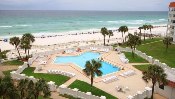 A pool area with a view of the beach and ocean, surrounded by palm trees and lounge chairs.