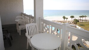 A balcony with white chairs and a table overlooking a beach with palm trees and a pool.