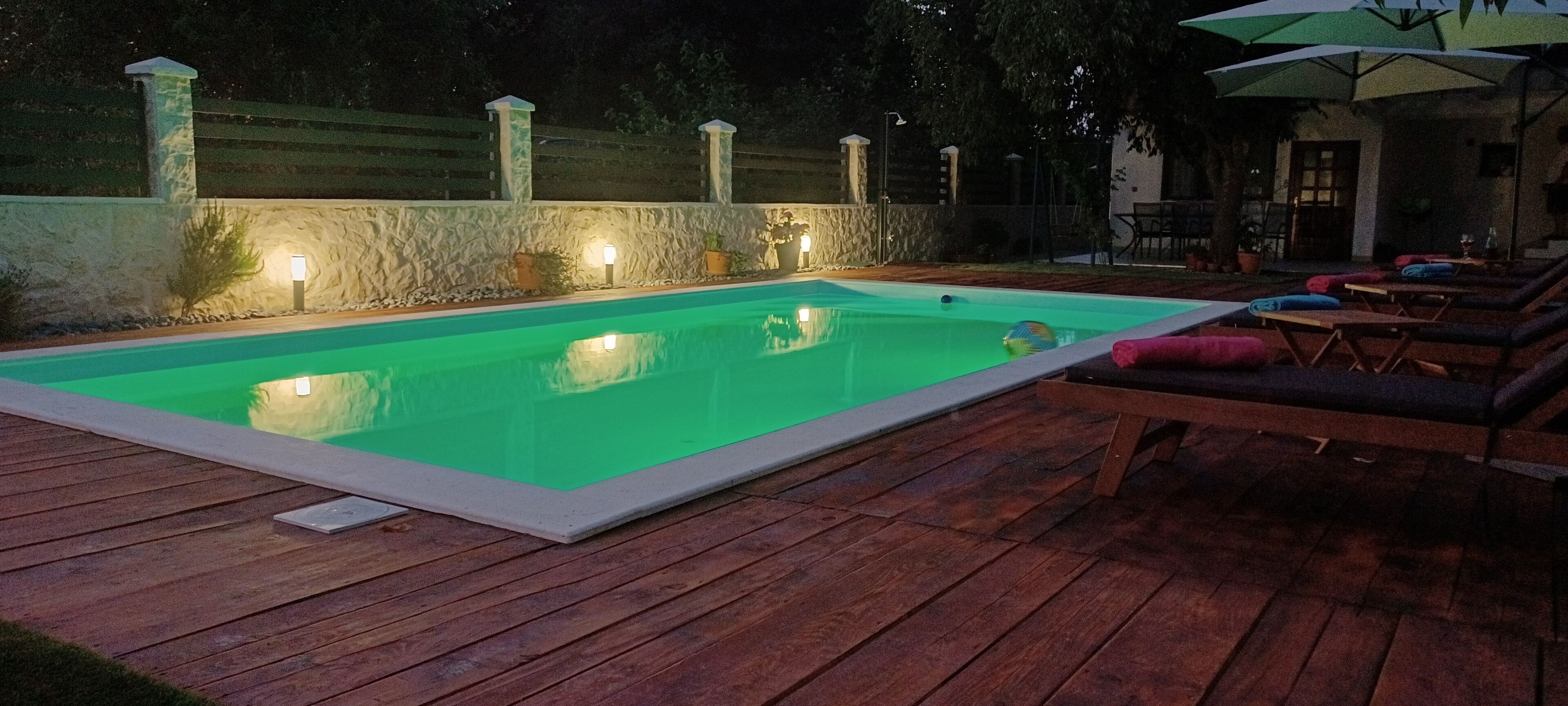 Ein rechteckiger Pool mit Steinmauer und Beleuchtung, umgeben von einer Holzterrasse und Liegestühlen.