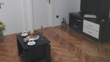 En stue med et sort sofabord, hvide døre, et fjernsyn på et sort stativ og et gulv med fiskebensmønster.