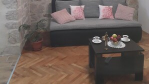 Una habitación acogedora con pared de piedra, un sofá gris con cojines rosa, suelo de madera y una pequeña mesa con una taza de café y fruta.