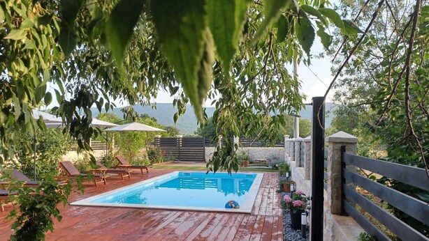 Una piscina rectangular con una terraza de madera, rodeada de césped y un muro de piedra.