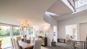 Une salle à manger spacieuse avec une grande table en bois, des chaises blanches et un lustre. De grandes fenêtres offrent une vue sur un jardin.
