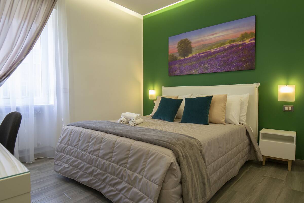 A neatly made bed with a beige quilt, two pillows, and a white headboard. There is a painting of a field with purple flowers on the wall above the bed.
