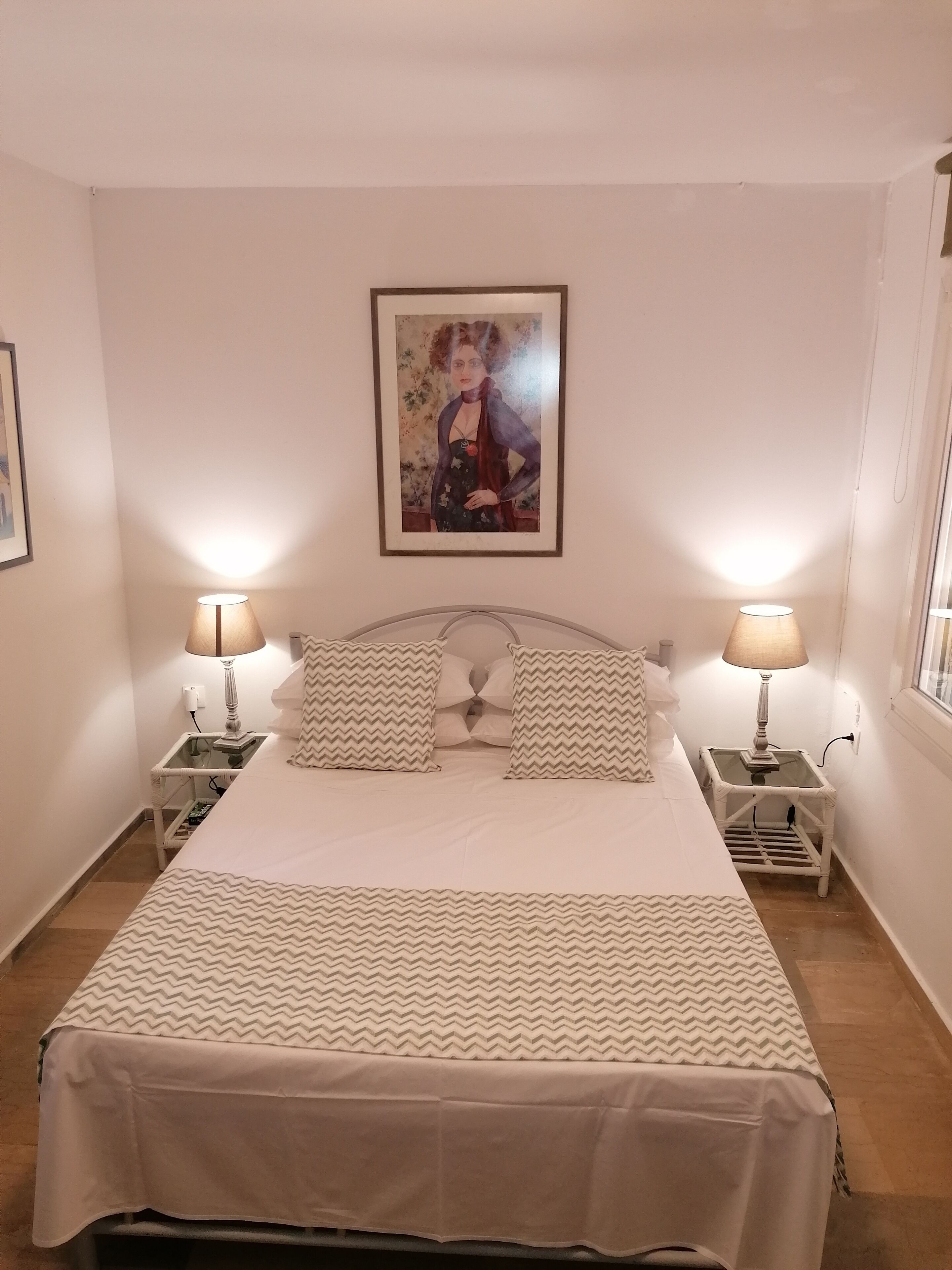 A neatly made bed with a patterned bedspread, two matching pillows, and two bedside lamps on white nightstands. A framed picture hangs above the bed.