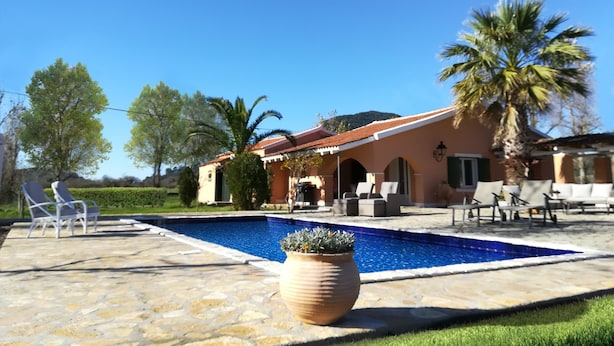 A pool area with lounge chairs, a stone patio, and a house with a red roof. - Corfu Golf Countryhouse (Corfu)