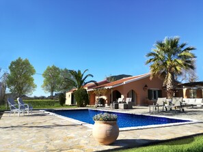 A pool area with lounge chairs, a stone patio, and a house with a red roof.