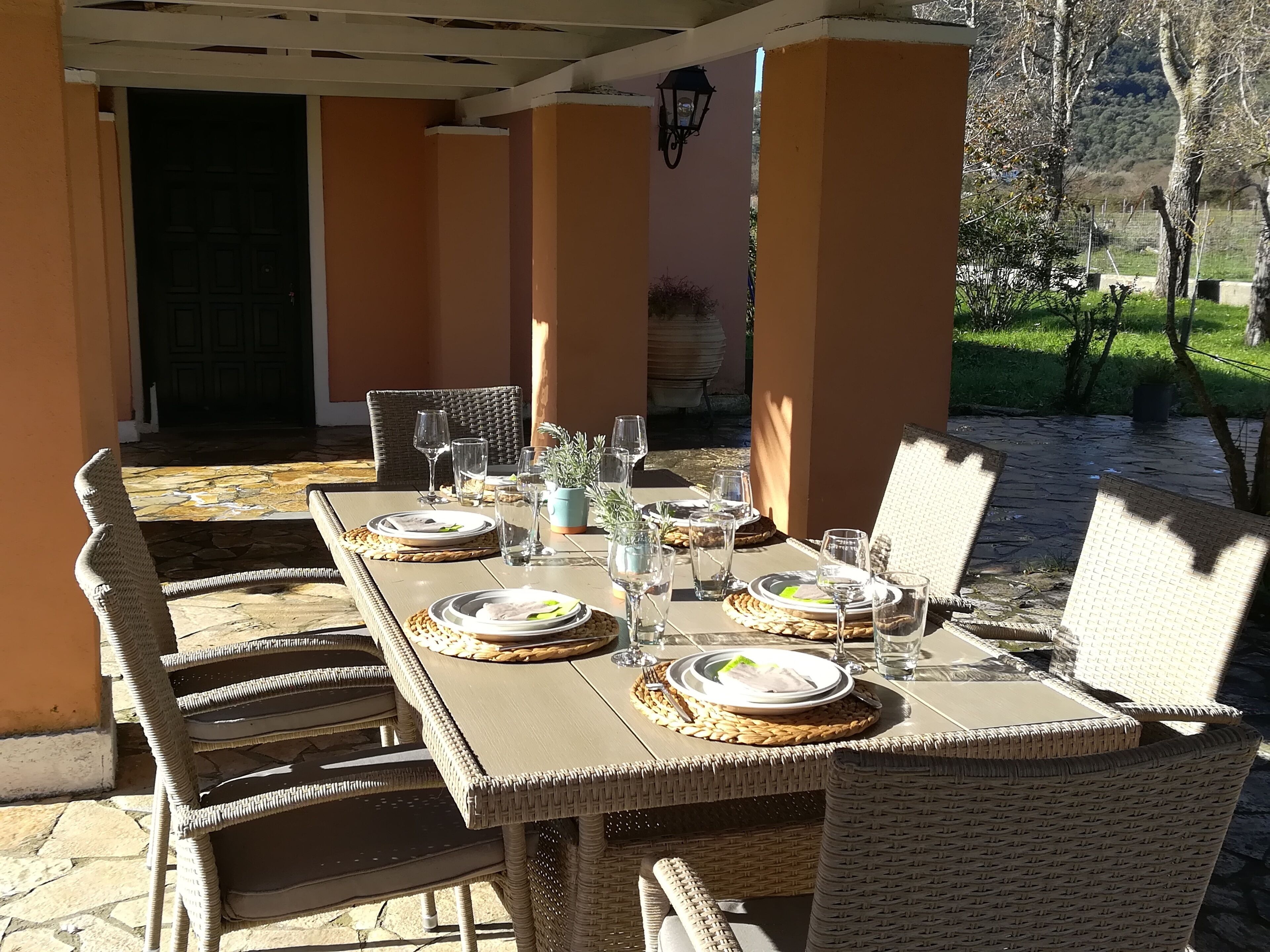 Un área de comedor al aire libre con una mesa preparada para cuatro, rodeada de sillas de mimbre, y vistas a un jardín y montañas al fondo.