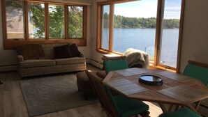 Quarto aconchegante com uma janela ampla de frente para um lago, uma mesa de madeira com cadeiras e um sofá com almofadas.