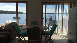 Uma área de refeições com mesa e cadeiras, janelas amplas com vista para um lago e barcos, e uma porta de vidro deslizante que dá para uma varanda externa.