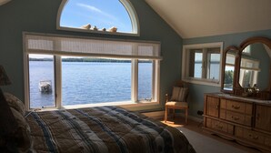 Quarto com uma grande janela com vista para um lago, uma cama com roupa de cama listrada, uma cadeira e uma cômoda.