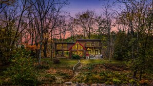 A two-story cabin with a red roof, surrounded by trees and a small stream. - River Time Cabin -riverfront, wifi, screen porch, 3 king beds, view, wood stove (Great Cacapon)