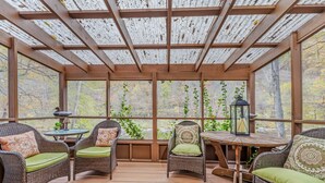 A covered outdoor seating area with wicker chairs and a table, surrounded by large windows offering views of trees and a body of water. - River Time Cabin -riverfront, wifi, screen porch, 3 king beds, view, wood stove (Great Cacapon)