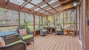 A covered outdoor deck with wicker furniture and a table, surrounded by glass panels and wooden beams. - River Time Cabin -riverfront, wifi, screen porch, 3 king beds, view, wood stove (Great Cacapon)