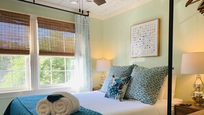 A neatly made bed with a teal quilt, two patterned pillows, and a rolled towel. There is a window with blinds, a wall-mounted lamp, and a framed artwork above the bed. - Stylish 2 BED, 2 BATH Shotgun-style house near beach! (Galveston)