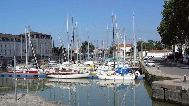 Un puerto deportivo con varias embarcaciones amarradas, un edificio de arquitectura clásica al fondo y un paseo pavimentado a orillas del agua.