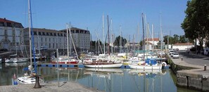 A marina with multiple boats docked, a building with classical architecture in the background, and a paved walkway along the water's edge.