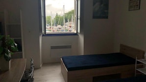 Una habitación pequeña y escasamente amoblada con una cama individual, un escritorio y una ventana con vista a un puerto deportivo.
