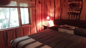 A cozy bedroom with a wooden bed, a window with a view of trees, and a lamp on a nightstand. - Cabaña Rústica Pucon, Maravillosa.  (Pucón)