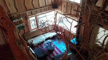 A cozy cabin interior with a wooden ceiling, a window with a view of trees, a fur-covered bench, and a blue rug.