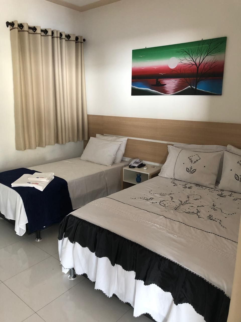 A hotel room with two beds, a nightstand, a painting, and a window with curtains.