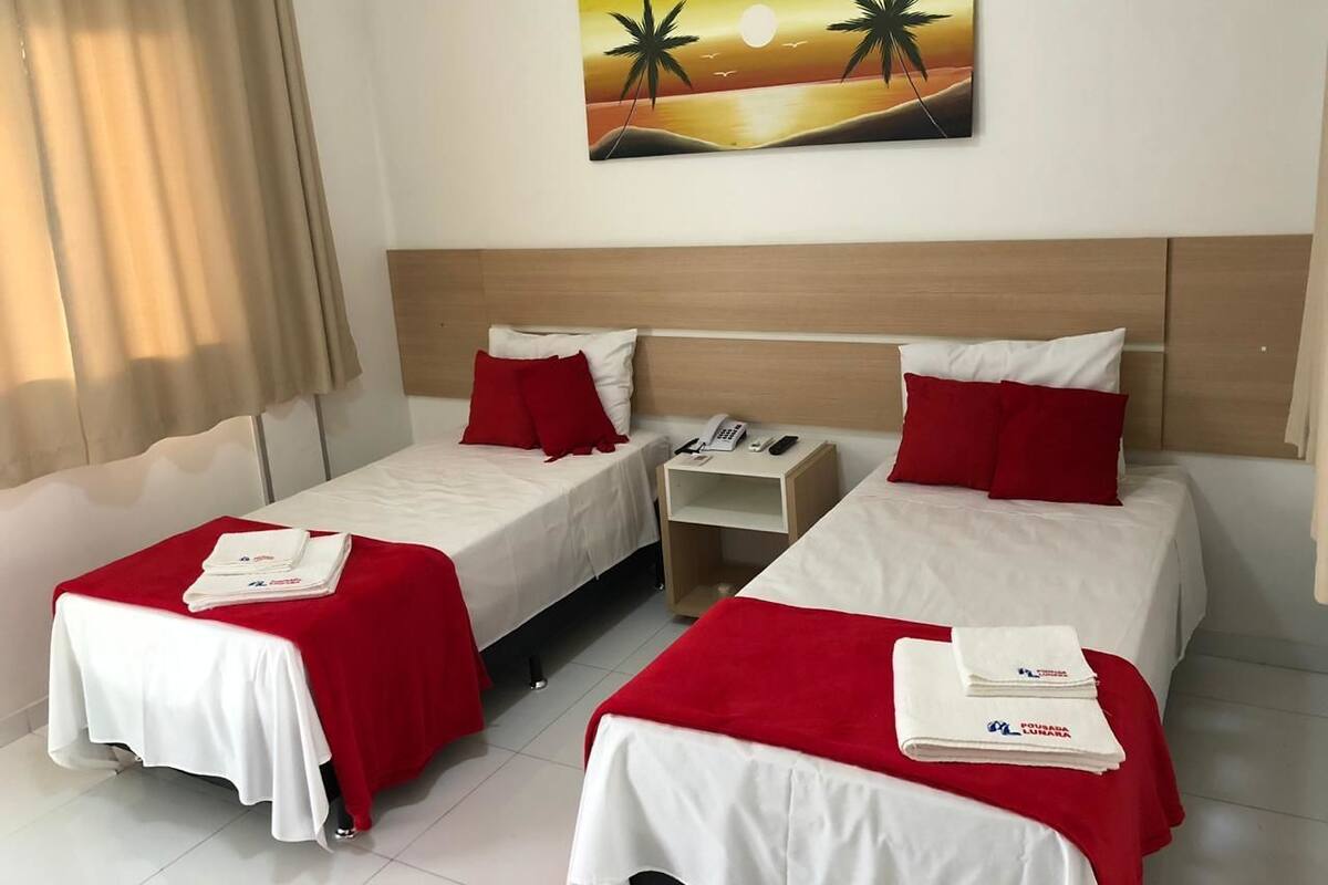 Ein Hotelzimmer mit zwei Betten, jedes mit einer roten Decke und einem weißen Kopfkissen, einem Nachttisch mit Telefon sowie einem Palmen-Gemälde an der Wand.